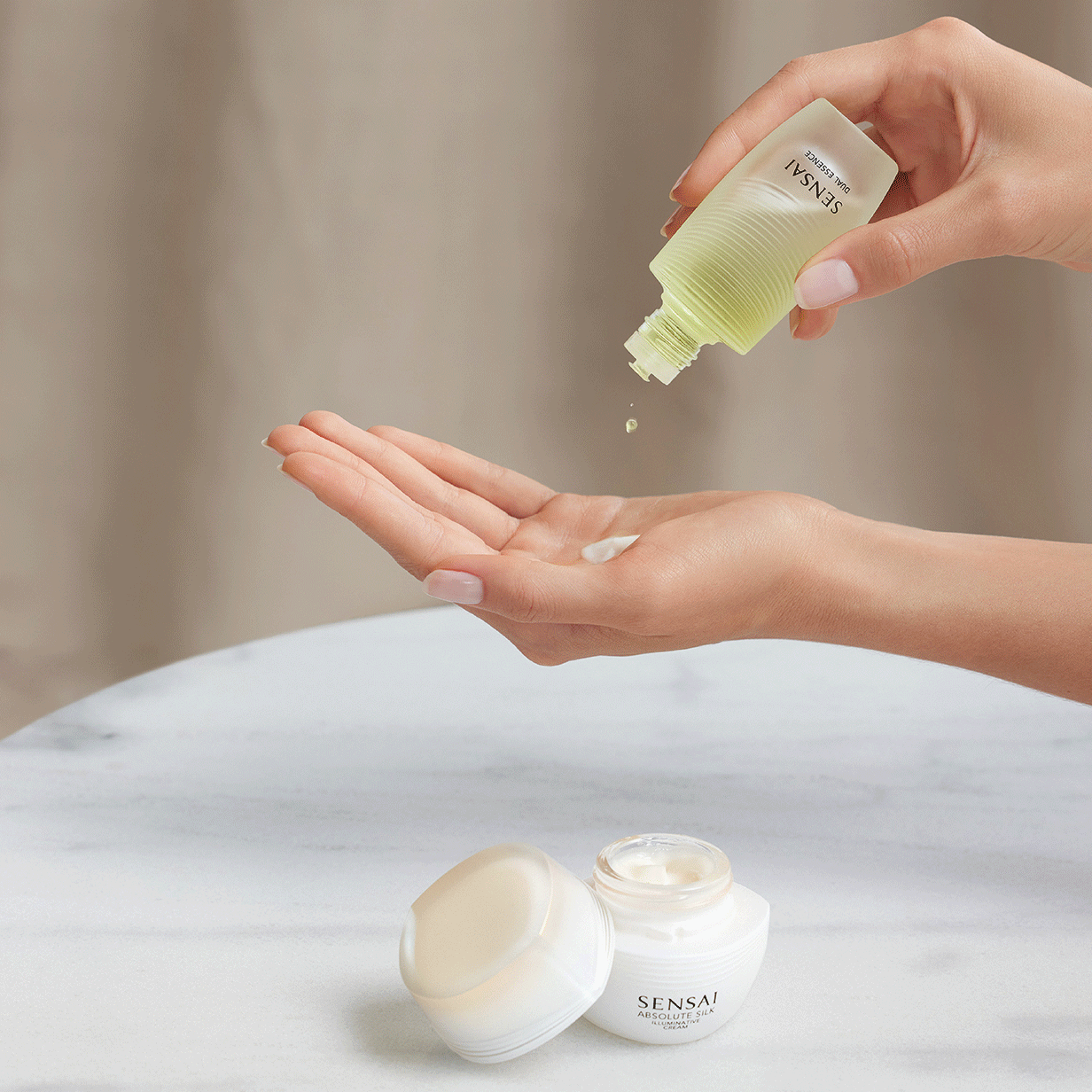 Absolute Silk Illuminative Cream e Dual Essence di SENSAI su un tavolo, formula della crema su un modello ed essenza sulla mano 